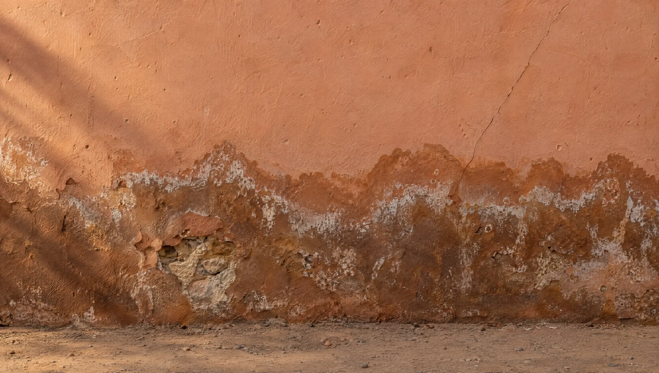 Base d'un mur en enduit ocre rongée par l'humidité : auréole sombre, salpêtre blanc, enduit décollé.